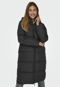 Manteau long noir rembourré avec capuche, doté d'une fermeture éclair et d'un design matelassé. Texture lisse et coupe ample offrent chaleur et confort.
