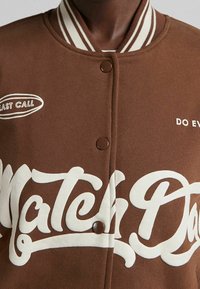 Brun varsityjacka med vit text. Har en ribbad krage och knappstängning. Anmärkningsvärda tryck: "LAST CALL" och "MATCH DAY".