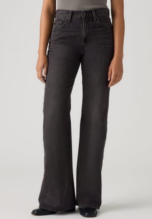 Femme portant un jean noir taille haute à jambes larges et des chaussures noires, debout sur un fond clair uni.