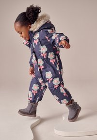 Combinezon impermeabil de iarnă, culoare bleumarin, cu modele florale în roz și verde. Prezintă un guler din blană artificială și manșete elastice, asortat cu cizme de iarnă gri.