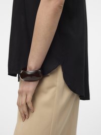 Henkilön käsivarsi, jossa musta yläosa ja beige housut, ranteessa paksu tummanruskea rannerengas.