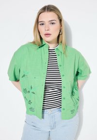 Camicetta verde a maniche corte con design floreale ricamato, abbinata a un top a righe bianche e nere. Jeans azzurri con perle applicate.