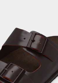 Σανδάλι από σκούρο καφέ δέρμα με δύο ρυθμιζόμενους ιμάντες με αγκράφες και ορατή πορτοκαλί ραφή κατά μήκος των άκρων.