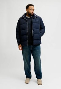 Navy puffer jacket with a zip collar, worn over a textured knit sweater, paired with light blue jeans and beige shoes.
