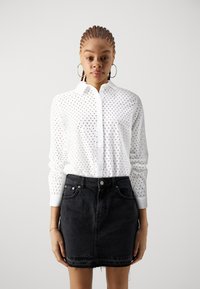 Camicia bianca con bottoni, dotata di un motivo ritagliato, maniche lunghe e colletto, abbinata a una minigonna in denim nera con orlo sfilacciato.