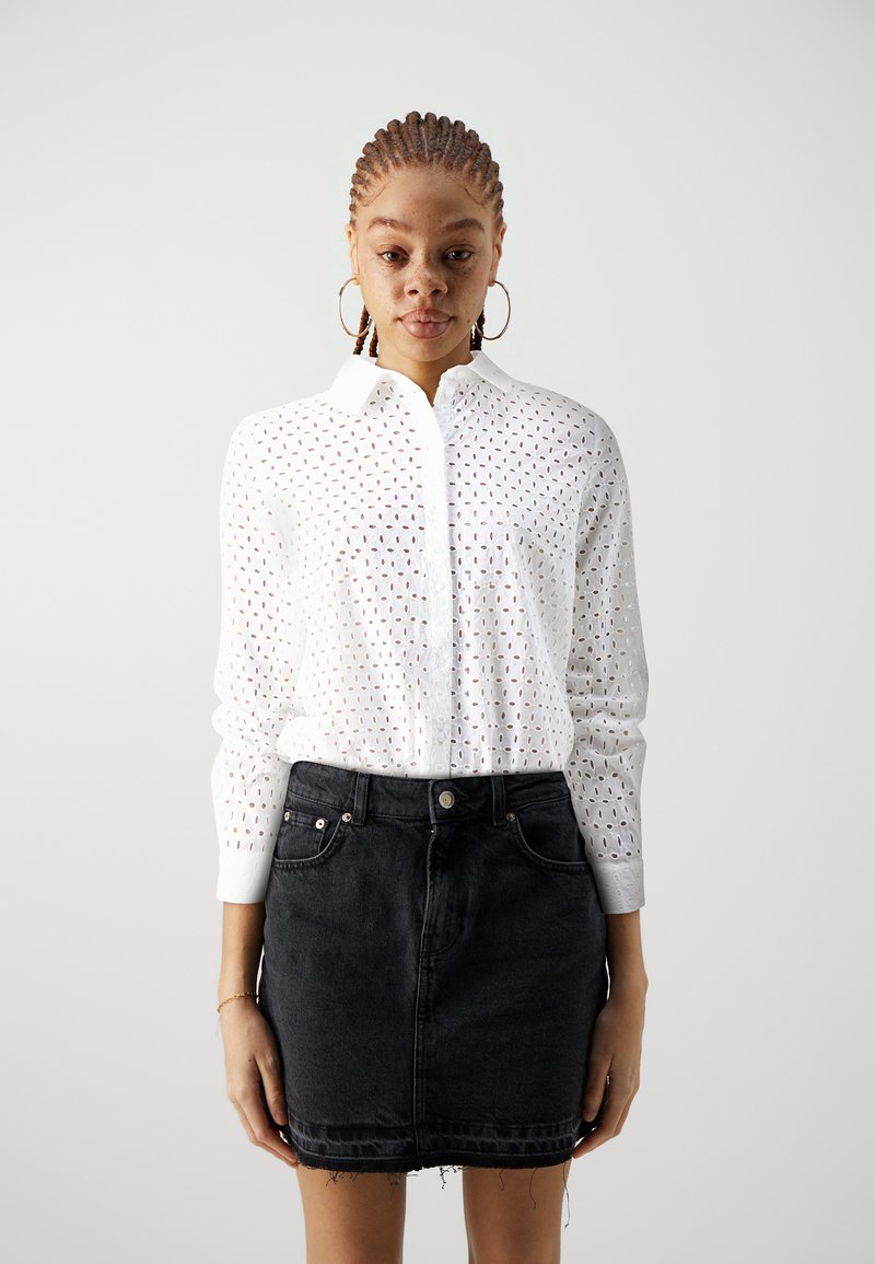 Camicia bianca con bottoni, dotata di un motivo ritagliato, maniche lunghe e colletto, abbinata a una minigonna in denim nera con orlo sfilacciato.