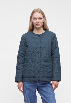 Jeune femme aux longs cheveux blonds portant une veste matelassée bleu foncé avec des boutons-pression et deux poches avant, associée à un jean bleu.