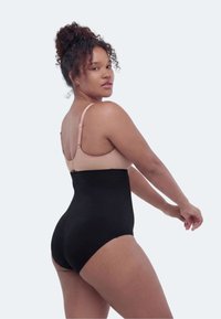Kvinne med krøllete hår i en hudfarget BH og svart formende undertøy med høyt liv, poserer med armene strukket bakover mot en ensfarget lys bakgrunn.