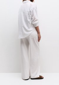 Witte linnen blouse met een ontspannen pasvorm en lange mouwen, gecombineerd met losse, lichtgekleurde broek, beide met minimale textuur en ontwerp.