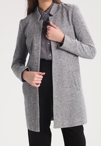 Cappotto grigio con texture, con colletto, maniche lunghe, apertura frontale e tasche laterali. Indossato sopra una camicia con bottoni e pantaloni neri.