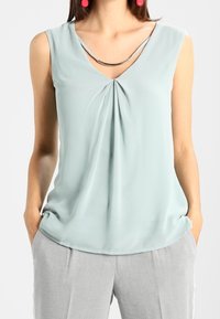 Kvinna som bär en ärmlös ljusgrön blus med framfåll och grå byxor, händerna i fickorna, röda runda örhängen och ett silverhalsband.