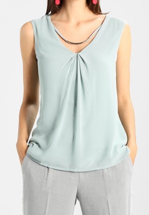 Femme portant un haut sans manches de couleur vert clair avec un pli sur le devant et un pantalon gris, les mains dans les poches, des boucles d'oreilles rondes rouges et un collier en argent.