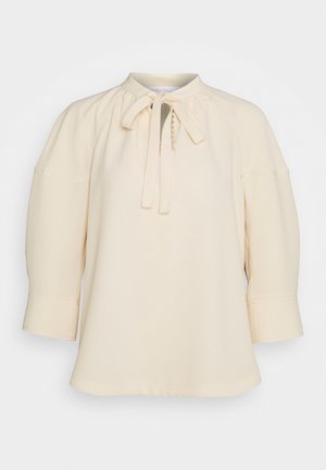 Blusa crema con collo alto, dettaglio a fiocco, maniche a tre quarti e sottili rifiniture cucite sulle maniche e sul corpino.