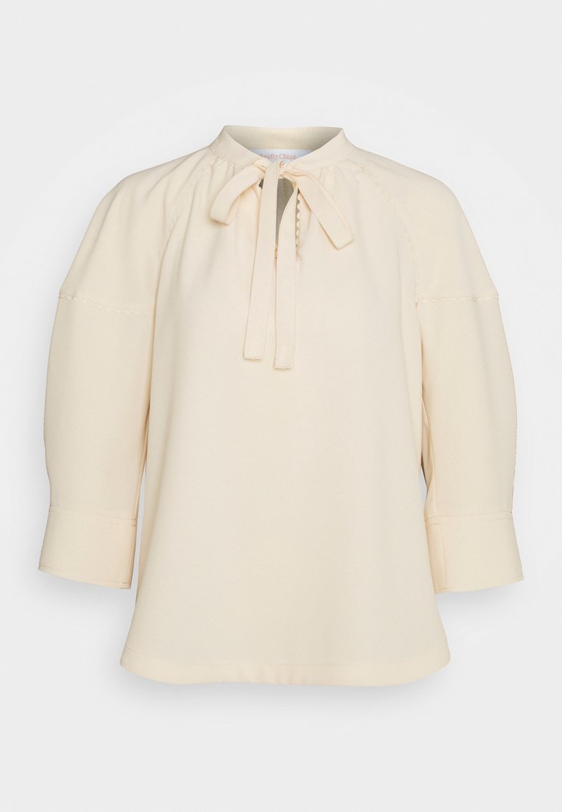 Blusa crema con collo alto, dettaglio a fiocco, maniche a tre quarti e sottili rifiniture cucite sulle maniche e sul corpino.