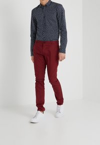 Chemise à manches longues à motif navy avec des designs géométriques blancs, associée à un pantalon bourgogne ajusté et des chaussures blanches, se tenant sur un fond uni.