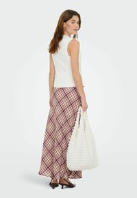 Haut blanc sans manches associé à une longue jupe à carreaux bordeaux et crème. Le modèle porte un sac blanc texturé et des chaussures à talons marron.