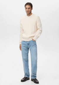 Maglione color crema a maglia intrecciata, jeans azzurri e scarpe nere. Il maglione presenta un motivo testurizzato, scollo rotondo e maniche aderenti.