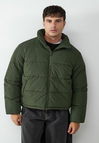 Grüne Pufferjacke mit Reißverschluss und hohem Kragen, die horizontale gesteppte Abschnitte und eine glatte, glänzende Stoffoberfläche aufweist.