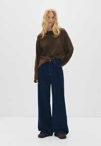 Hnědá oversized košile s dlouhým rukávem v kombinaci s širokými tmavě modrými džínami a hnědými nazouvacími botami. Jednoduchý, uvolněný střih s ležérním designem.