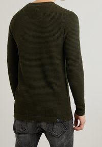 Maglione a maniche lunghe in maglia waffle verde oliva con un motivo testurizzato, dettagli di cucitura minimali e un piccolo etichetta con logo vicino all'orlo.