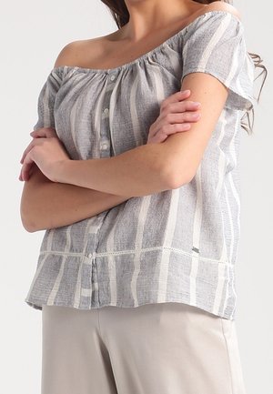 Kvinde iført off-shoulder, kortærmet, grå og hvide stribet bluse med knapper, armene krydset, og lyse bukser.