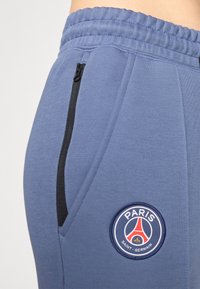 Blå joggebukser med elastisk midjebånd, med en sidelomme med glidelås og et rundt Paris Saint-Germain-logo i rødt, hvitt og blått.