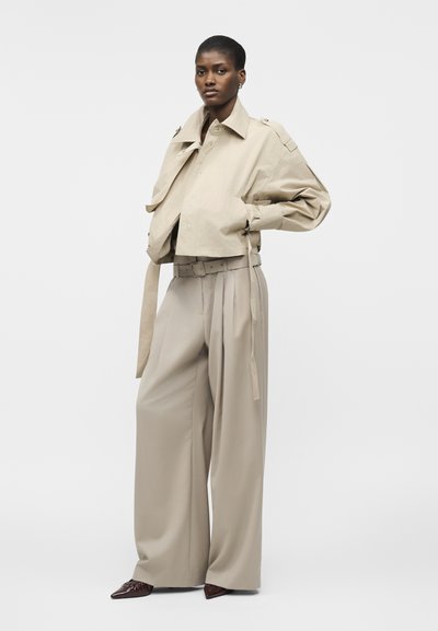 Γυναίκα στέκεται φορώντας μπεζ oversized μπουφάν και φαρδιά πλισέ παντελόνια με ζώνη, σε συνδυασμό με σκούρα μυτερά παπούτσια, πάνω σε απλό φόντο.