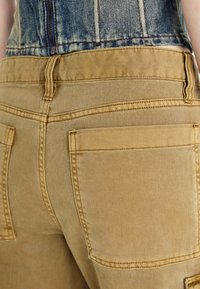 Khaki džíny šortky s viditelnými zadními kapsami, akcenty v šití a mírně vybledlým povrchem, navržené s hladkou texturou a přiléhavým střihem.