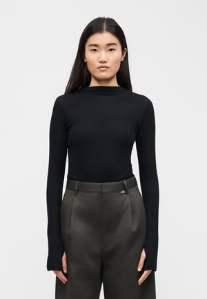 Kvinde med langt mørkt hår, iført en tætsiddende sort langærmet top og mørkegrå bukser med høj talje, stående mod en ensartet hvid baggrund.