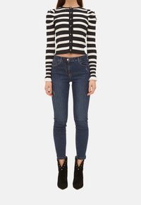 Žena v černo-bílém vodorovně pruhovaném zkráceném topu na knoflíky, modrých skinny džínách a černých botách na podpatku se cvočky, stojící.