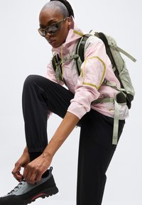 Άτομο που φοράει γυαλιά ηλίου, ροζ μπουφάν και ανοιχτό πράσινο σακίδιο πλάτης δένει μαύρο παπούτσι πεζοπορίας, ντυμένο για υπαίθρια δραστηριότητα.