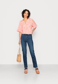 Mulher com cabelo encaracolado, vestindo uma blusa floral rosa, jeans azuis, saltos laranja e azul marinho, segurando uma bolsa de palha trançada, em pé contra um fundo branco.