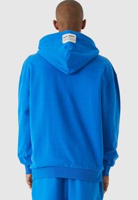 Felpa blu realizzata in morbido tessuto, dotata di un ampio cappuccio, polsini e orlo a coste, e un'etichetta sul collo. Texture liscia e vestibilità comoda.