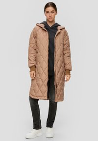 Beige gewatteerde lange jas met capuchon, voorzien van een ritssluiting en knoopaccenten. Gecombineerd met een grijze top, donkere jeans en witte sneakers.
