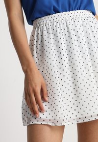 Bílá sukně s černými puntíky, elastický pas, lehký materiál a splývavý střih. Viditelná ruka na pase.