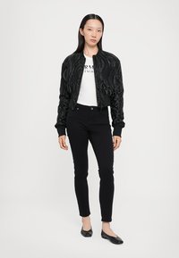Giacca bomber nera con una texture ondulata, polsini a coste, chiusura zip, abbinata a una t-shirt bianca con grafica e jeans skinny neri.