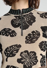 Suéter beige con patrones de hojas negras, cuello alto con diseño a rayas y cierre frontal con cremallera plateada.