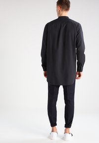 Camisa negra de manga larga hecha de tela suave, con un cuello clásico y un dobladillo curvado. Combinada con pantalones oscuros de cuadros y zapatos blancos.