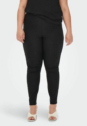 Vrouw staand, gekleed in nauwsluitende zwarte broek en witte open-toe sandalen met hak, tegen een effen lichte achtergrond.