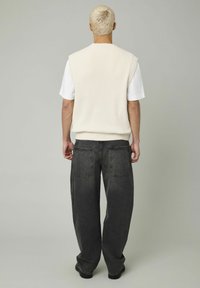 Pull sans manches en tricot blanc cassé porté sur un t-shirt blanc, associé à un pantalon en denim gris foncé ample avec des poches arrière.