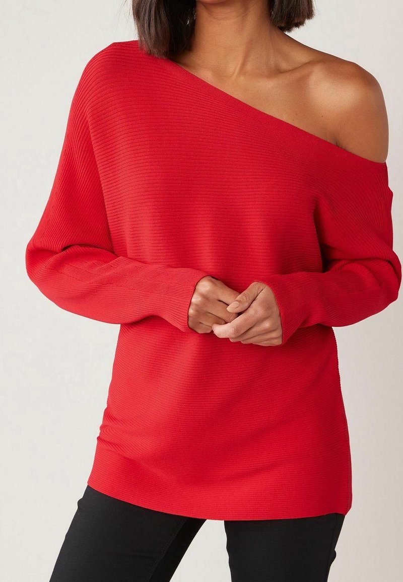 Femme portant un pull rouge côtelé à épaules dénudées avec manches longues et pantalon noir, mains jointes devant.