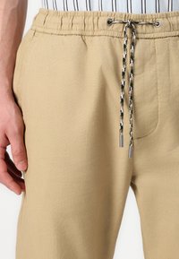 Beige Jogginghose mit elastischem Bund, aus strukturiertem Stoff und mit kontrastierenden, geflochtenen Kordeln mit Metallelementen.