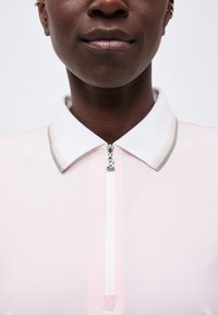 Nærbillede af en person iført en lyserosa skjorte med hvid krave og en frontlynlås med en "B" trækflig.