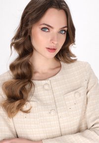 Giacca beige testurizzata con due bottoni rotondi, due tasche frontali e leggeri luccichii. Tessuto liscio con una vestibilità su misura e design senza colletto.