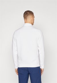 Pullover bianco con colletto alto, caratterizzato da un motivo a strisce verticali testurizzate, maniche lunghe e orlo dritto. Indossato con pantaloni blu.