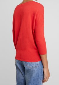 Pull tricot rouge avec manches côtelées 3/4 de longueur et encolure arrondie. Texture lisse, avec une couture au centre du dos.