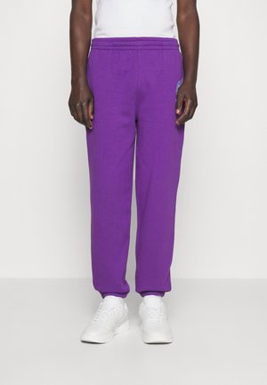 Personne portant un pantalon de survêtement violet vif associé à des baskets blanches et un haut sans manches blanc, se tenant devant un fond uni.