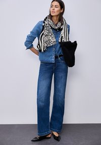 Femme portant une chemise en denim bleu et un jean, une écharpe imprimé animal et rayée, tenant un sac noir, debout sur un sol gris avec un mur blanc en arrière-plan.