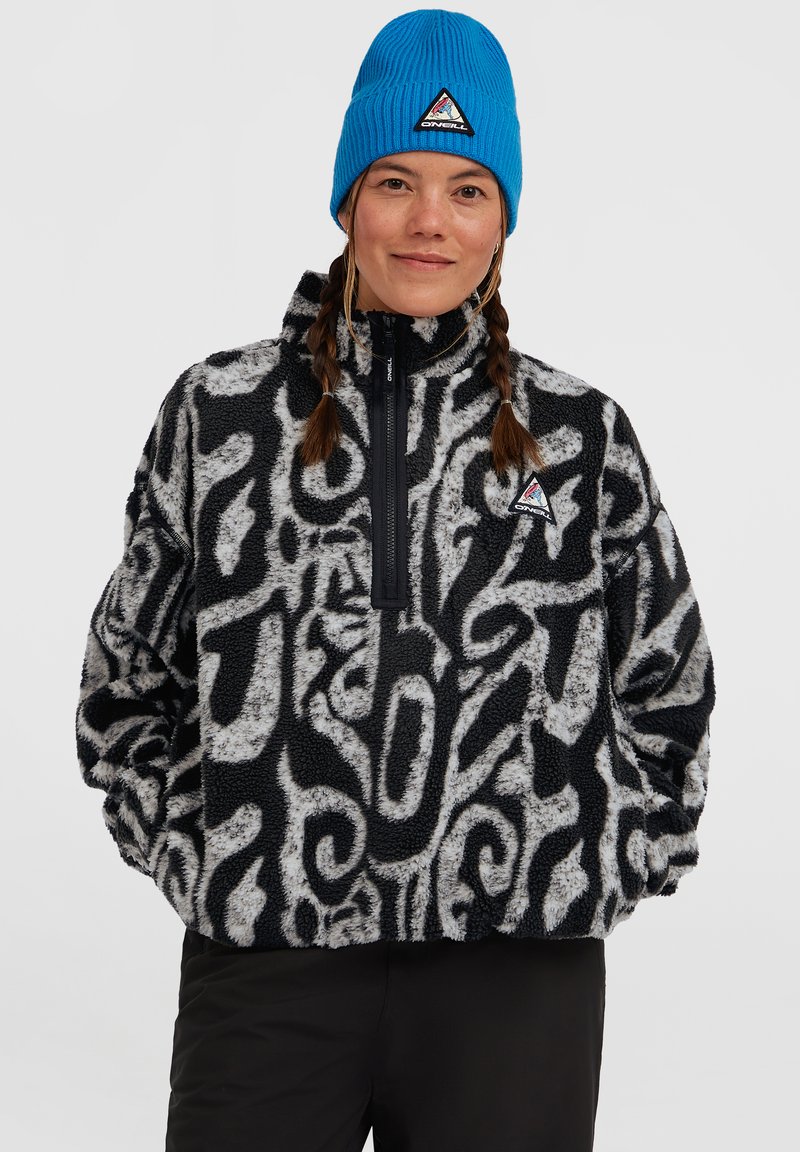 Vrouw met gevlochten haar die een zwart-wit gestreepte fleecejack en een felblauwe muts draagt, tegen een effen achtergrond.