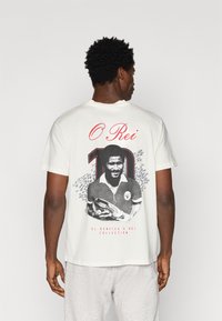 Λευκό βαμβακερό T-shirt με μαύρη και κόκκινη γραφική εκτύπωση ενός ποδοσφαιριστή με τον αριθμό "10" και το κείμενο "O Rei" στην πλάτη.